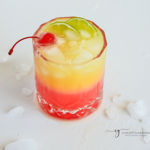 a highball glass with a layered red and yellow malibu sunset drink. Garnished with a cherry and lime slices.