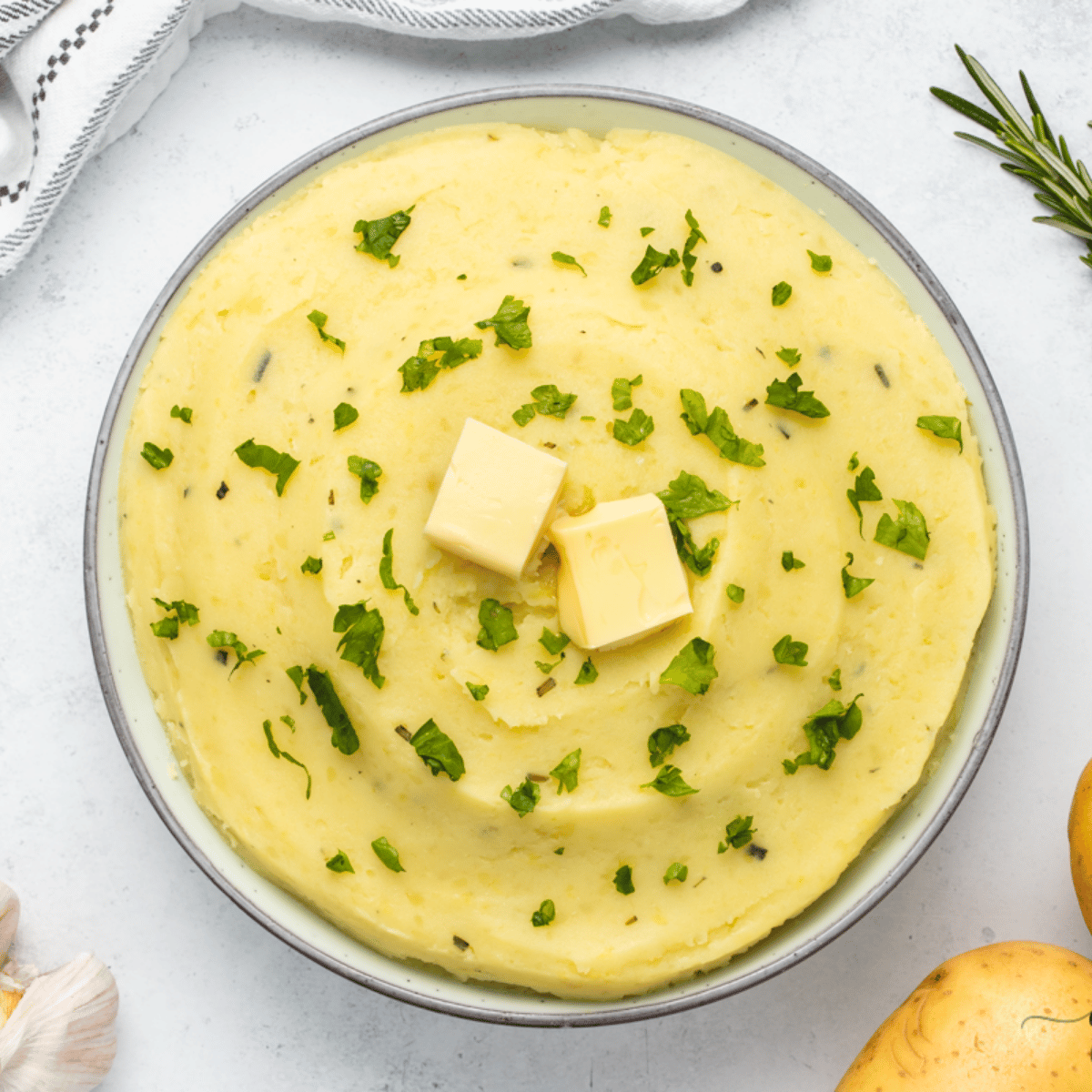 upclose shot of yellow mashed potatoes in a white bowl with two slabs of butter on top and green herbs