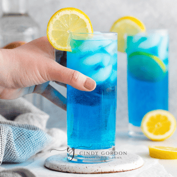 two clear tall glasses filled with ice and a blue liquid, topped with lemon slices, lemon slices also on counter, vodka bottle in back and towel to left side sex on the driveway drink