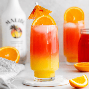 Sex on the beach drink malibu - 2 tall clear glasses filled with half moon ice, orange liquid on the bottom, red liquid on top and orange wheel on very top. Malibu rum bottle in the back. Orange slices on table