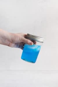 hand shaking a ball jar with lid. Inside blue liquid and ice cubes