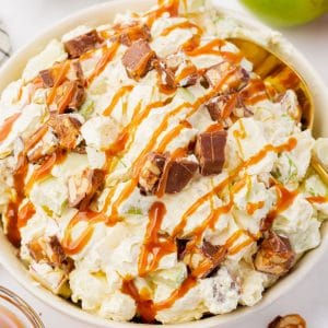 white bowl filled with fully white stuff, apple chunks snicker chunks and caramel syrup. On the side is red and green apples