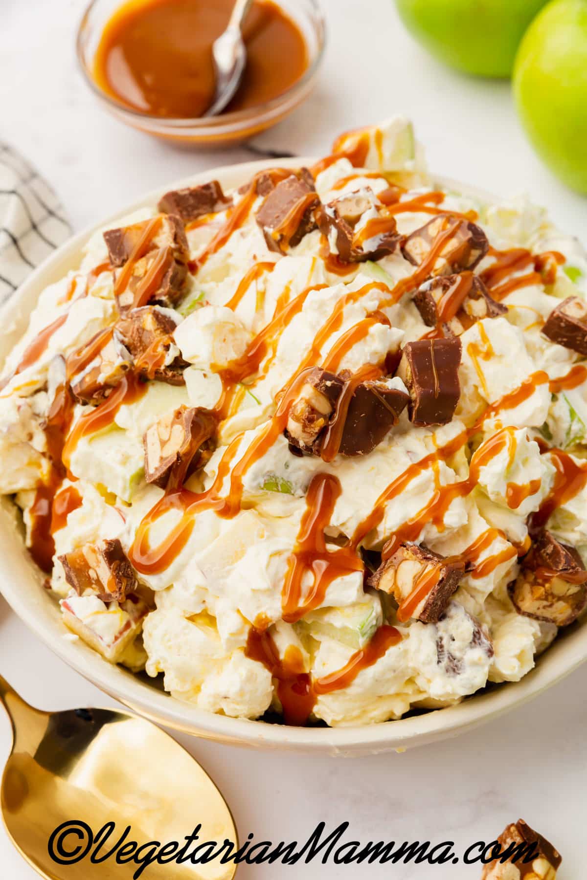 A white bowl filled with a mixture of white items, including apple chunks, Snickers chunks, and caramel syrup. On the side is red and green apples