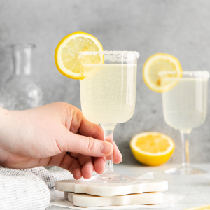 two clear glasses filed with off white/yellow liquid with lemon wheel on top. Lemon wheels on surface and grey cloth to left, a hand holding one of the glasses