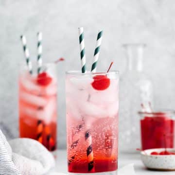 Two tall, clear glasses filled with red liquid at the bottom, clear fizzy liquid at the top with ice cubes and two striped straws in each glass. Each glass also has a cherry in it.