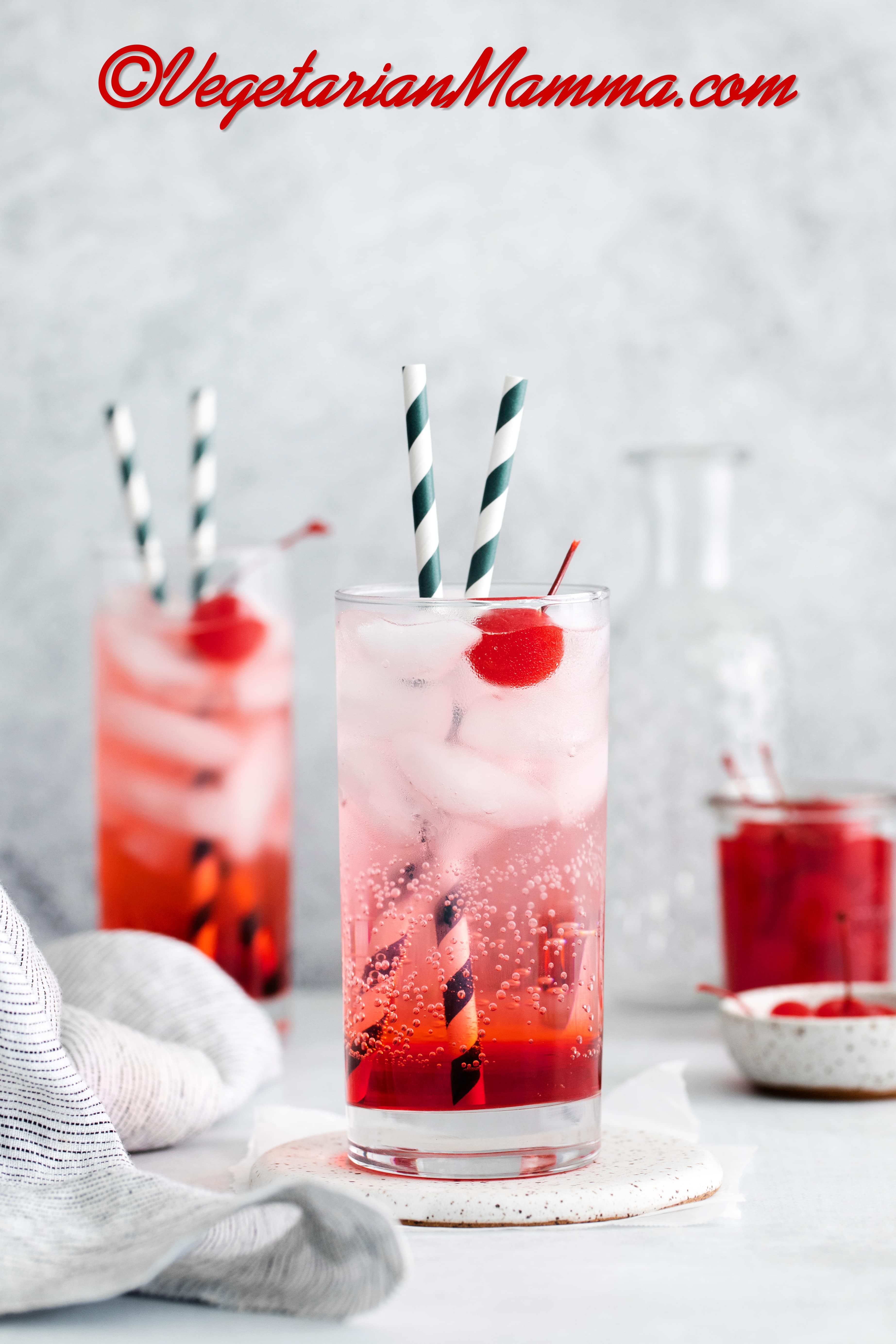 Two tall, clear glasses filled with red liquid at the bottom, clear fizzy liquid at the top with ice cubes and two striped straws in each glass. Each glass also has a cherry in it.
