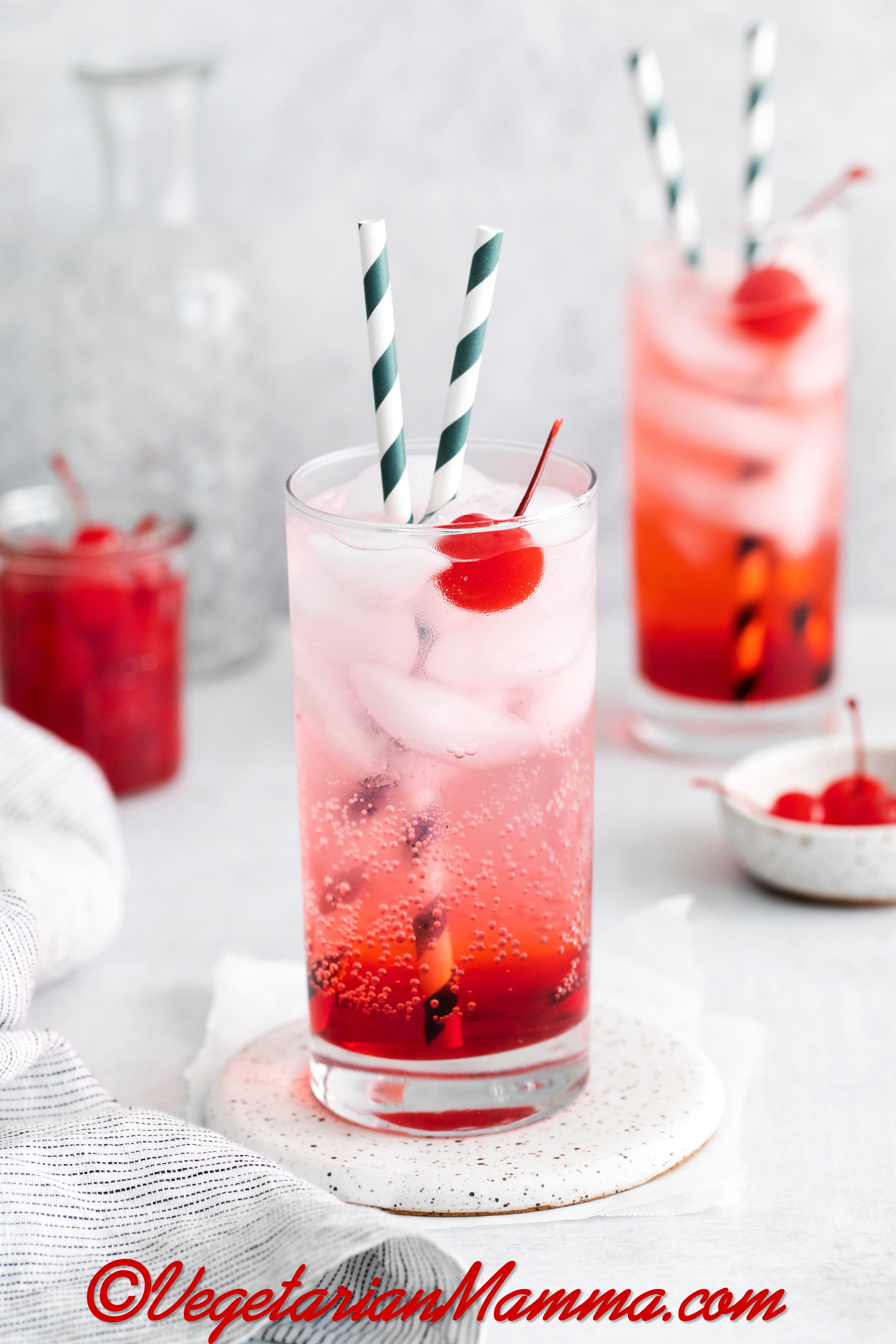 Two tall, clear glasses filled with red liquid at the bottom, clear fizzy liquid at the top with ice cubes and two striped straws in each glass. Each glass also has a cherry in it.