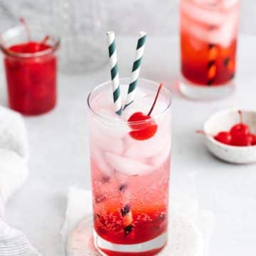 Two tall, clear glasses filled with red liquid at the bottom, clear fizzy liquid at the top with ice cubes and two striped straws in each glass. Each glass also has a cherry in it.