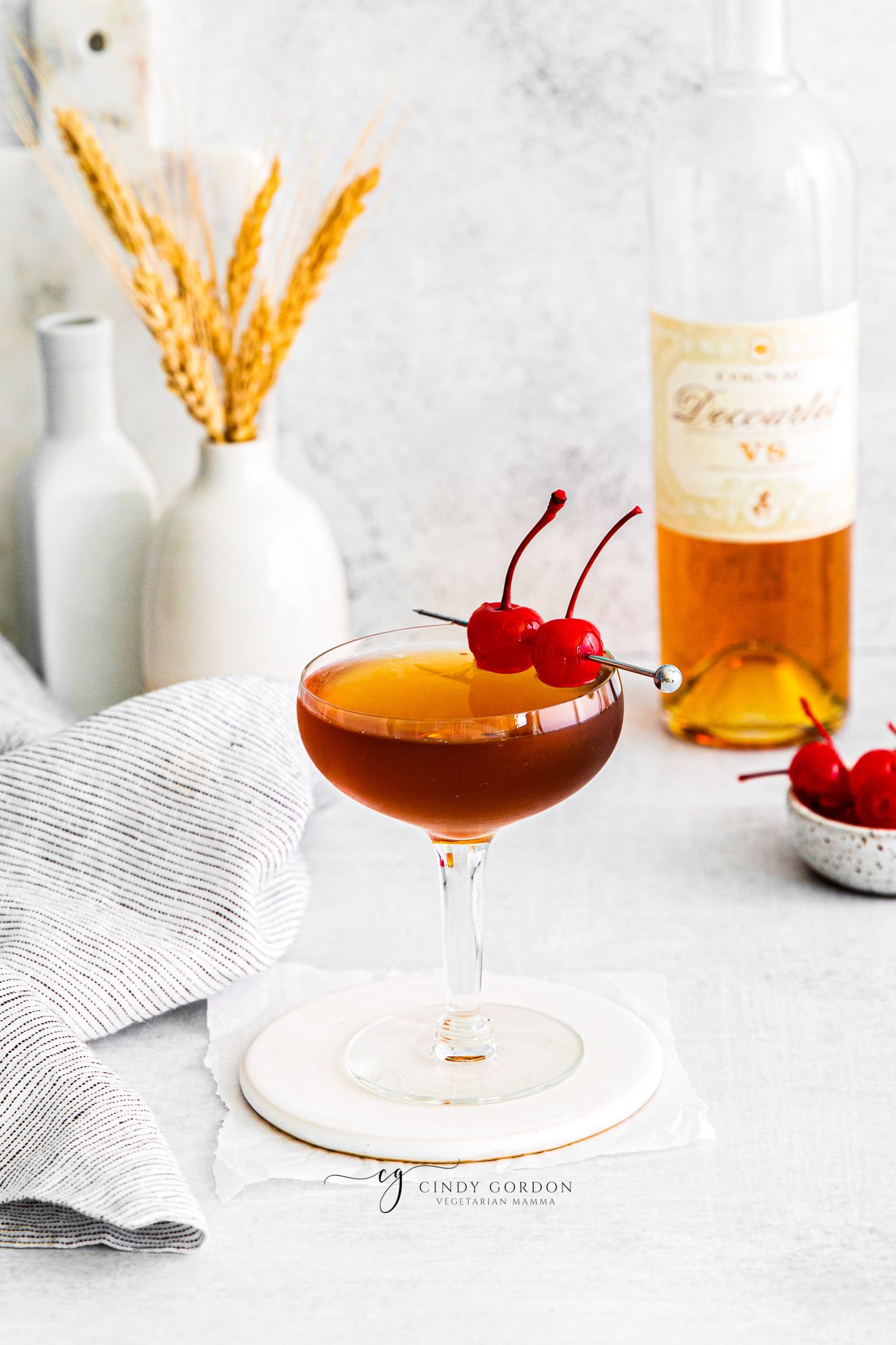 Pictured is a brandy manhattan. One glass full of orange and brown liquid with two cherries on stop. a bottle of brandy in the background with some cherries on a plate.