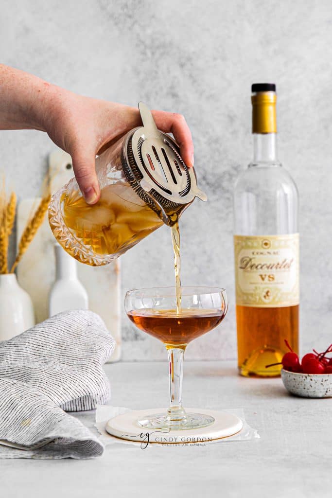 hand pouring brown liquid from a strainer into a glass, brandy bottle in back right