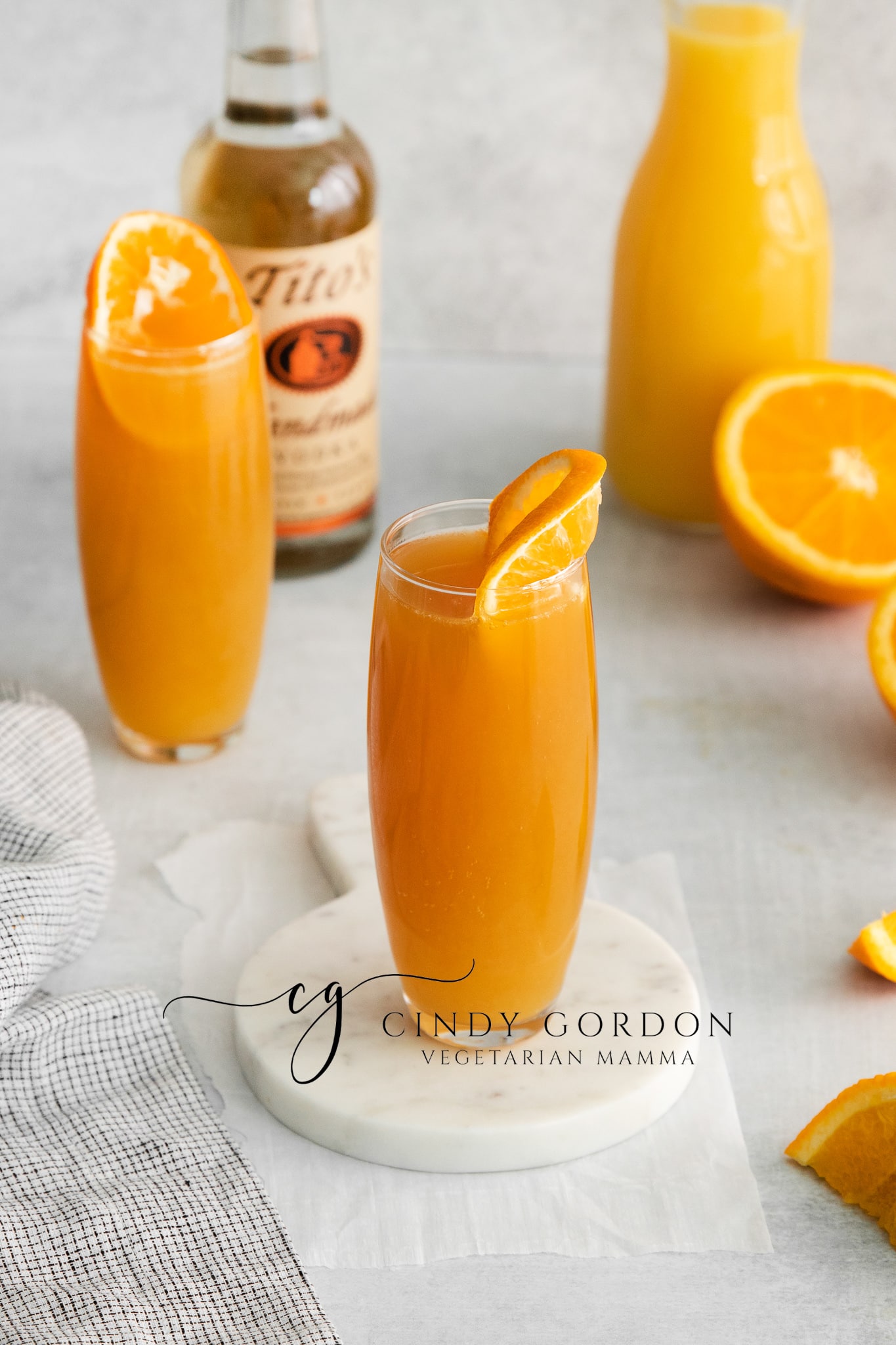 two tall clear glasses filled with orange liquid and topped with an orange slice. cut orange in back ground with a vodka bottle and orange juice jar