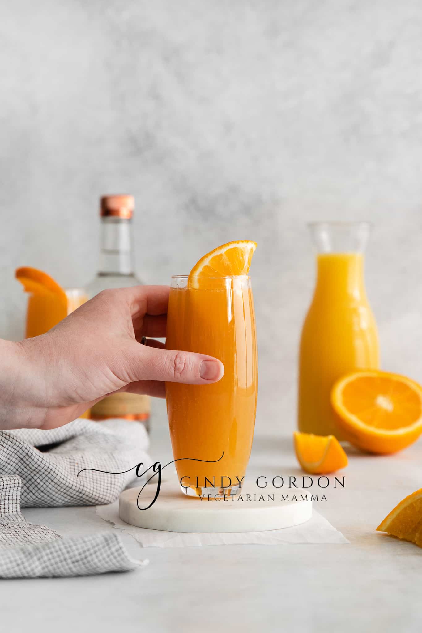 two tall clear glasses filled with orange liquid and topped with an orange slice. cut orange in back ground with a vodka bottle and orange juice jar. hand holding of the orange juice glasses