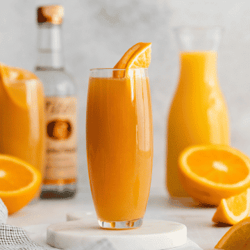 two tall clear glasses filled with orange liquid and topped with an orange slice. cut orange in back ground with a vodka bottle and orange juice jar