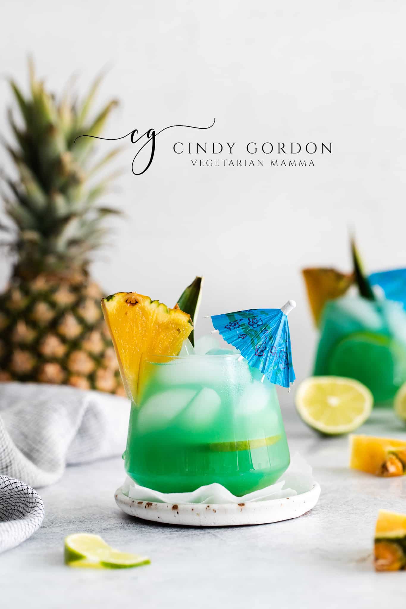 Pictured is two short glasses of mermaid water drink. The glasses are full of a blue green liquid with ice cubes. There is a pineapple chunk on the side of the glasses. A large pineapple in the background and lime slices on the table also a small blue drink umbrella in the forward most drink