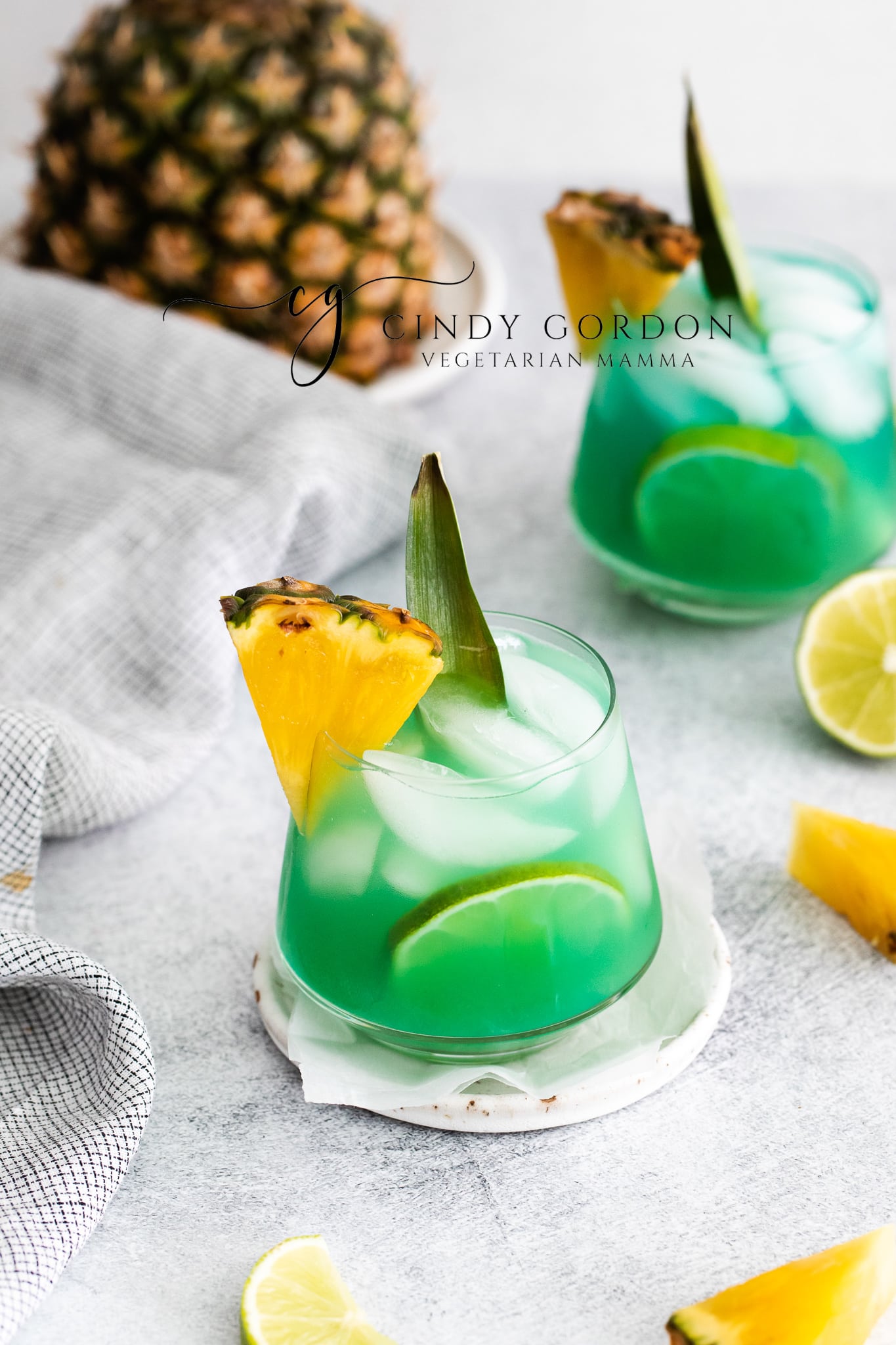 Pictured is two short glasses of mermaid water drink. The glasses are full of a blue green liquid with ice cubes. There is a pineapple chunk on the side of the glasses. A large pineapple in the background and lime slices on the table 