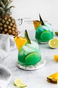 Pictured is two short glasses of mermaid water drink. The glasses are full of a blue green liquid with ice cubes. There is a pineapple chunk on the side of the glasses. A large pineapple in the background and lime slices on the table