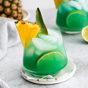 Pictured is two short glasses of mermaid water drink. The glasses are full of a blue green liquid with ice cubes. There is a pineapple chunk on the side of the glasses. A large pineapple in the background and lime slices on the table