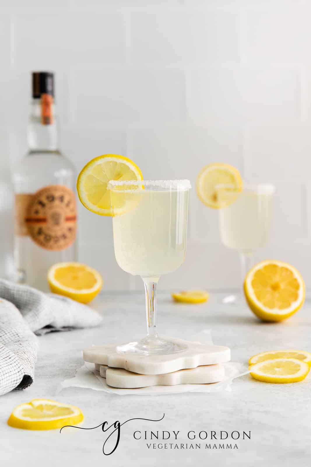 two clear glasses filed with off white/yellow liquid with lemon wheel on top. Lemon wheels on surface and grey cloth to left with vodka bottle in back left