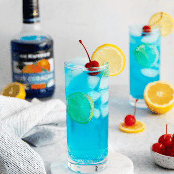 blue motorcycle drink - blue liquid with white ice cubes in a tall clear glass. cherry and lemon wheel on top and a bottle of blue alcohol in back
