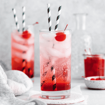 two tall clear glasses filled with red liquid at bottom, clear fizzy liquid at top with ice cubes and two striped straws in each glass. Each glass also has a cherry in it.