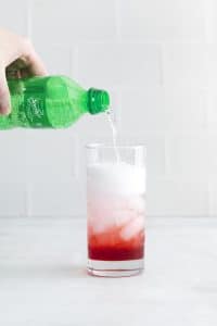 tall glass with red liquid and ice cubes sprite being poured in to glass