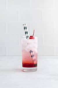 one tall clear glass filled with red liquid at bottom, clear fizzy liquid at top with ice cubes and two striped straws in glass. glass also has a cherry in it.