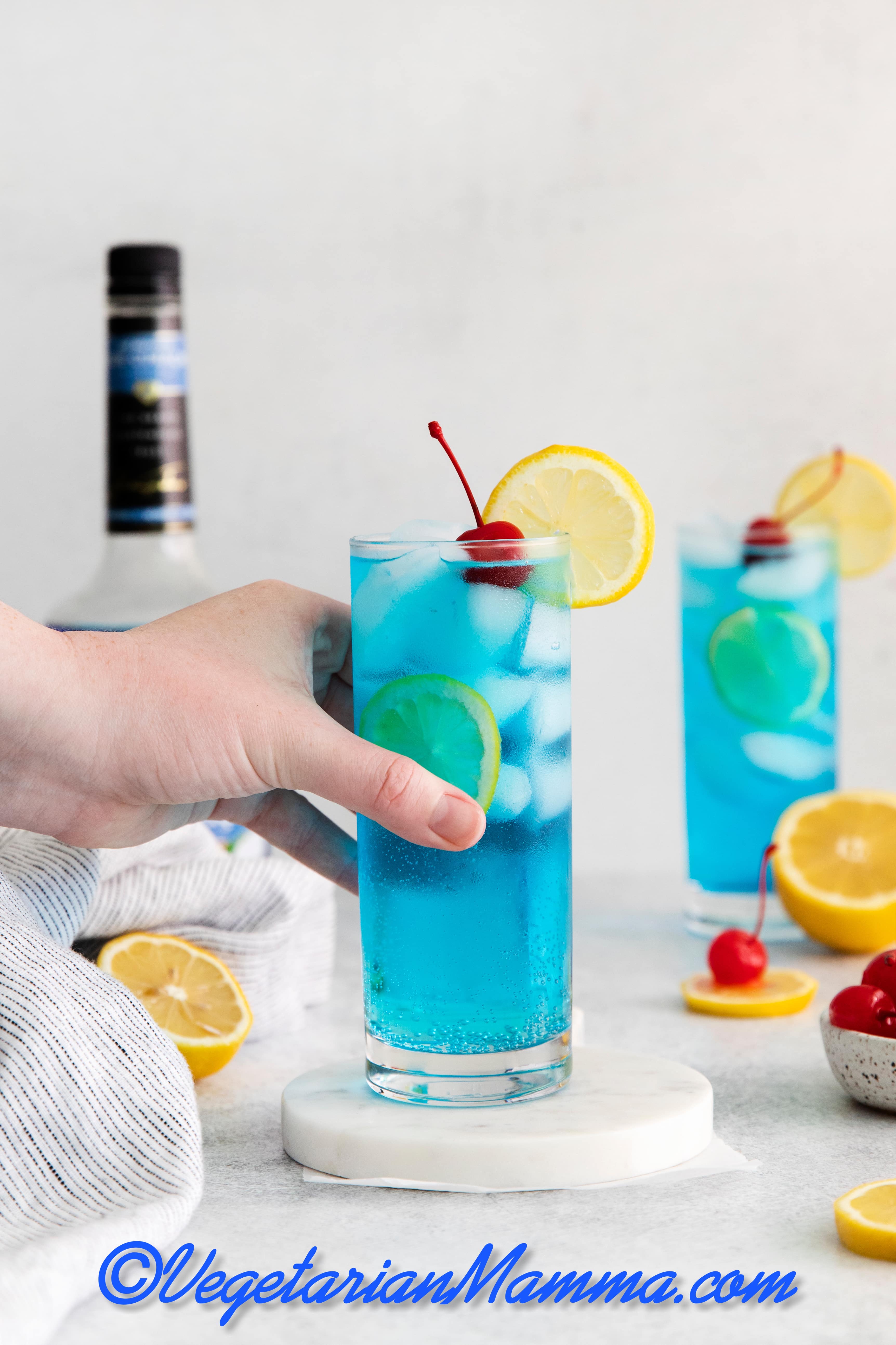 hand holding blue motorcycle drink - blue liquid with white ice cubes in a tall clear glass. cherry and lemon wheel on top