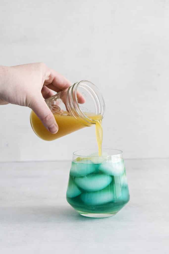 pineapple juice being poured into a glass with blue liquid and ice cubes
