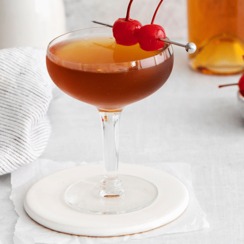 Pictured is a brandy manhattan. One glass full of orange and brown liquid with two cherries on stop. a bottle of brandy in the background with some cherries on a plate.