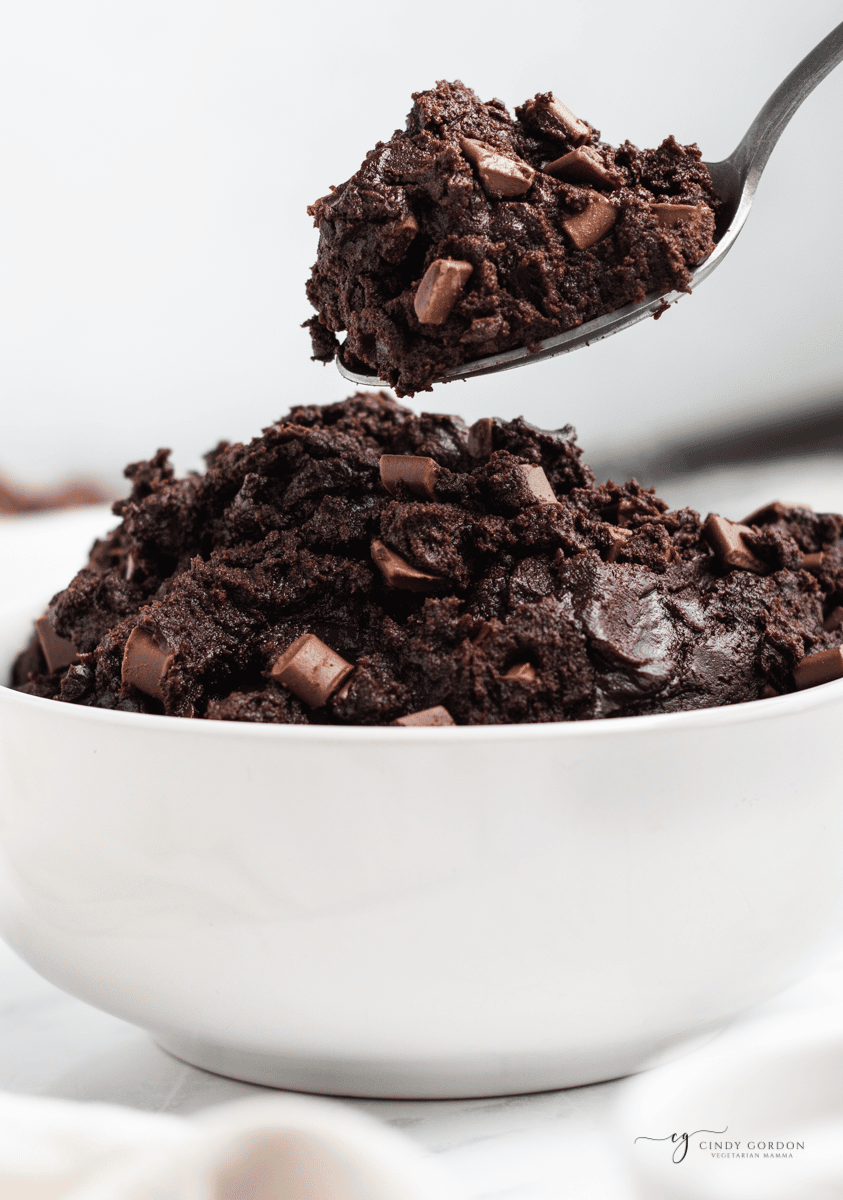 a white bowl filled with dark chocolate edible brownie batter. A spoon is lifting out a spoon full of it.