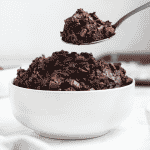 a white bowl filled with dark chocolate edible brownie batter. A spoon is lifting out a spoon full of it.