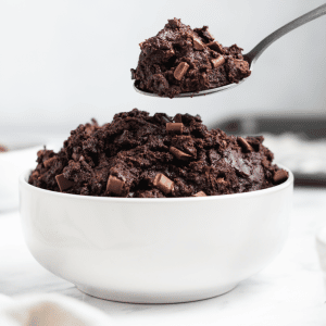 a white bowl filled with dark chocolate edible brownie batter. A spoon is lifting out a spoon full of it.