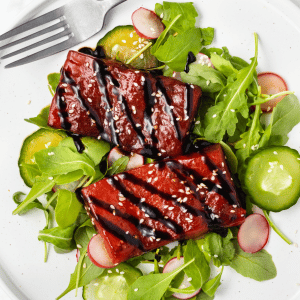 A white dinner plate of fresh greens topped with two rectangular watermelon steaks drizzled with balsamic glaze and sesame seeds