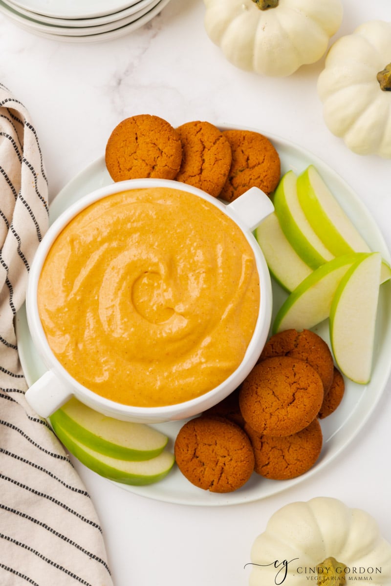 orange creamy pumpkin dip in a bowl, set on a plate of gingersnaps and sliced green apples.