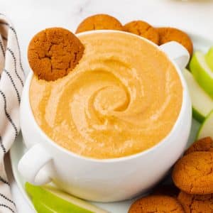 A large bowl with two handles, filled with creamy pumpkin dip. A gingersnap is in the bowl of dip. Under the bowl is a white plate filled with more gingersnaps and sliced green apples.