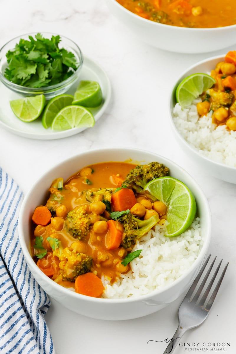 a bowl of pumpkin curry and rice on a table. In the background is a plate of lime wedges and cilantro. 