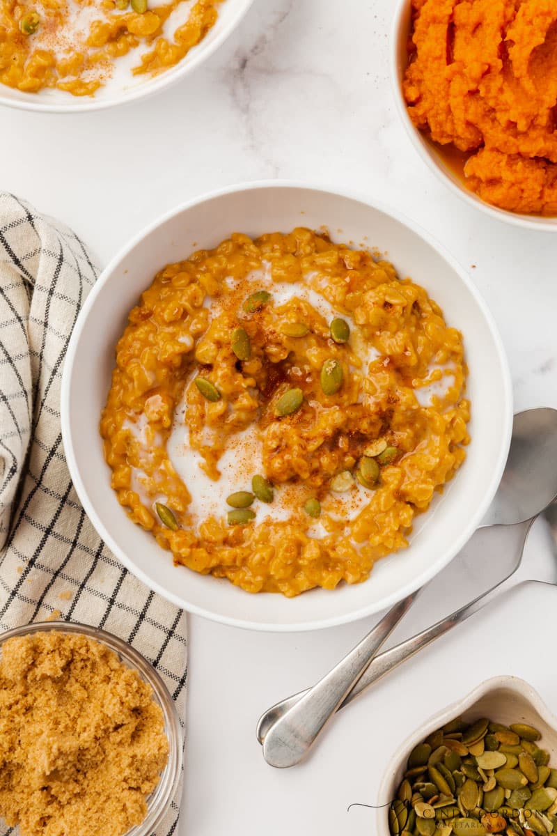 A white bowl filled with pumpkin oatmeal, topped with pepitas. Around the bowl is other bowls of the ingredients used to make the oatmeal.