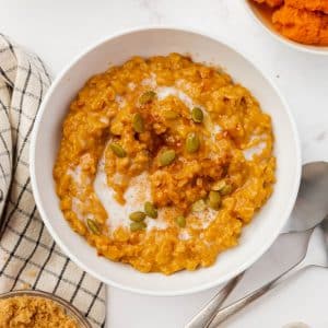 A white bowl filled with pumpkin oatmeal, topped with pepitas. Around the bowl is other bowls of the ingredients used to make the oatmeal.