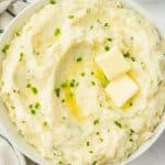 closeup of a bowl of mashed potatoes with chives and two pats of butter on top.