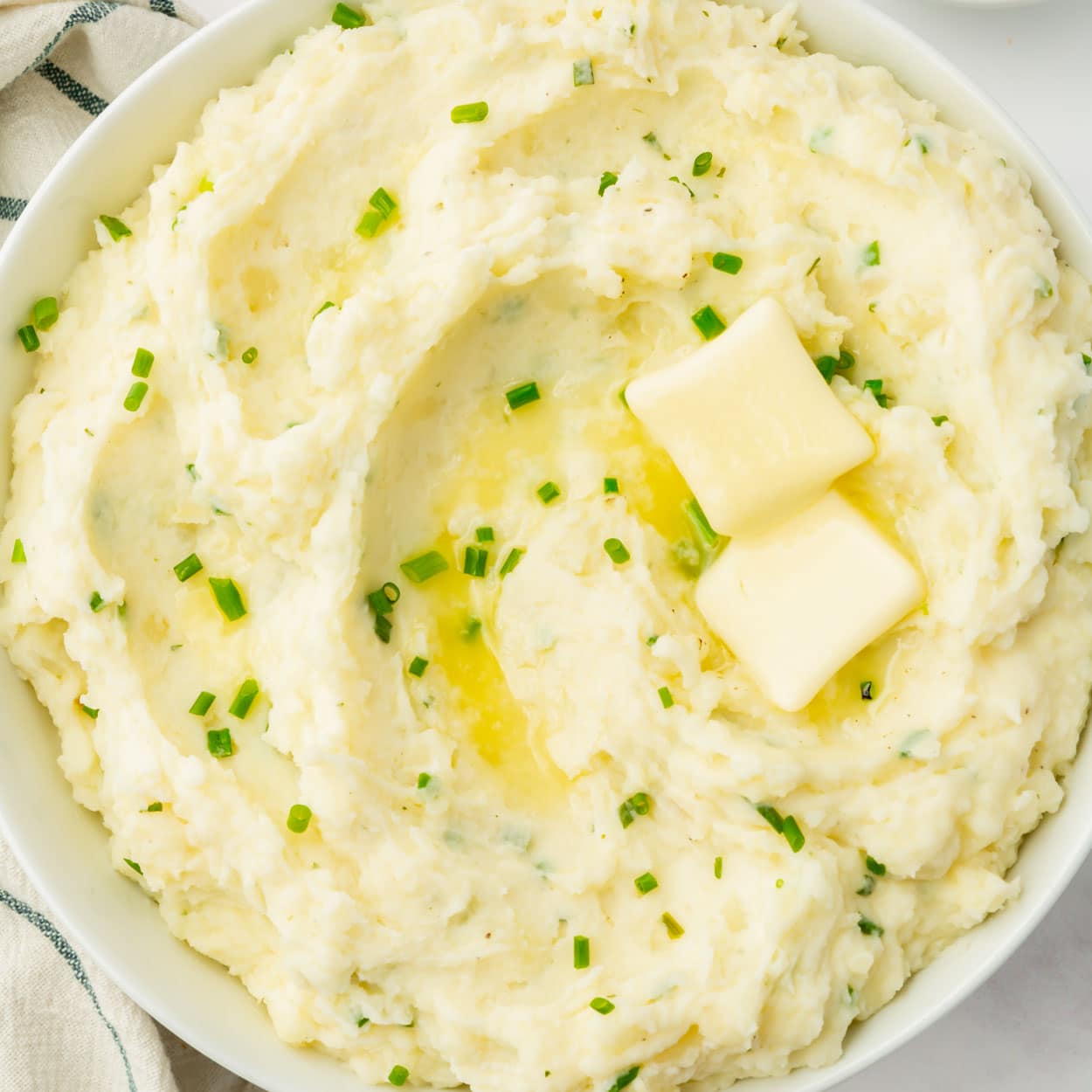 closeup of a bowl of mashed potatoes with chives and two pats of butter on top.