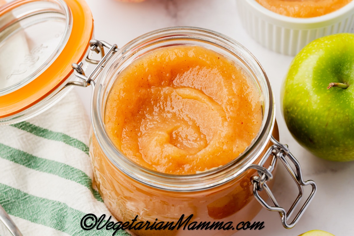 A lidded glass jar filled with homemade unsweetened applesauce. Around the jar are fresh apples. 
