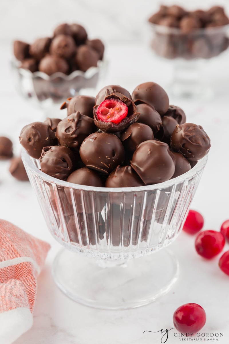 a footed glass dessert dish filled with chocolate covered cranberries. One has been bitten into to show the inside. 