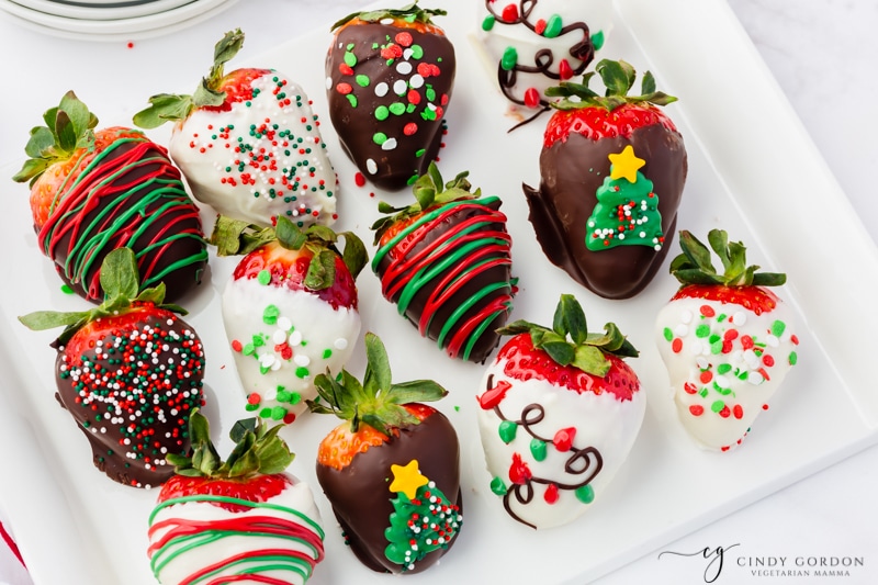 11 strawberries on a square platter. Each is dipped in milk or white chocolate. Each has a different christmas decoration on it with sprinkles and red and green candy melts. 