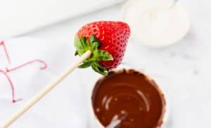 a strawberry on a skewer, being moved toward a bowl of melted chocolate