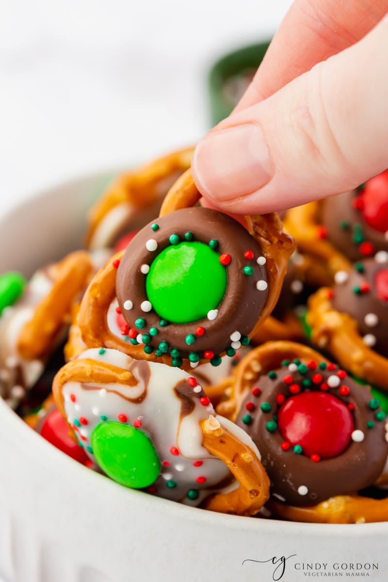 a hand picking up a pretzel covered in chocolate with holiday m&ms and sprinkles on it, removing it from a bowl of christmas pretzels.