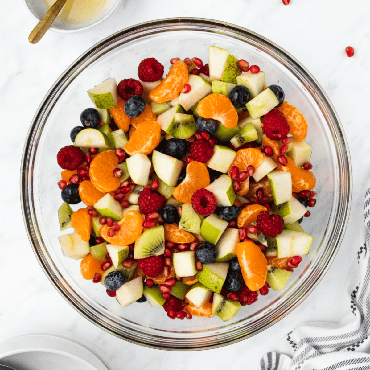 a clear glass bowl filled with fruit salad featuring clementines, green apples, and pomegranate seeds