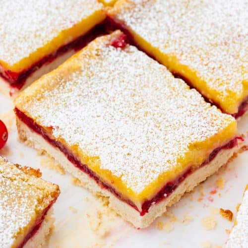 a large rectangle lemon bar with cranberry filling, dusted with powdered sugar, next to other squares of cranberry lemon bars.