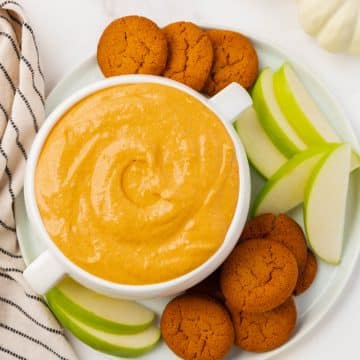 A large bowl with two handles, filled with creamy pumpkin dip. A gingersnap is in the bowl of dip. Under the bowl is a white plate filled with more gingersnaps and sliced green apples.