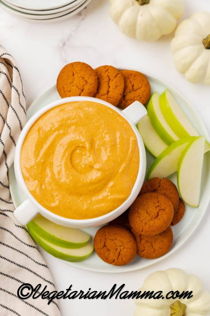 A large bowl with two handles, filled with creamy pumpkin dip. A gingersnap is in the bowl of dip. Under the bowl is a white plate filled with more gingersnaps and sliced green apples.