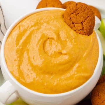 A large bowl with two handles, filled with creamy pumpkin dip. A gingersnap is in the bowl of dip. Under the bowl is a white plate filled with more gingersnaps and sliced green apples.